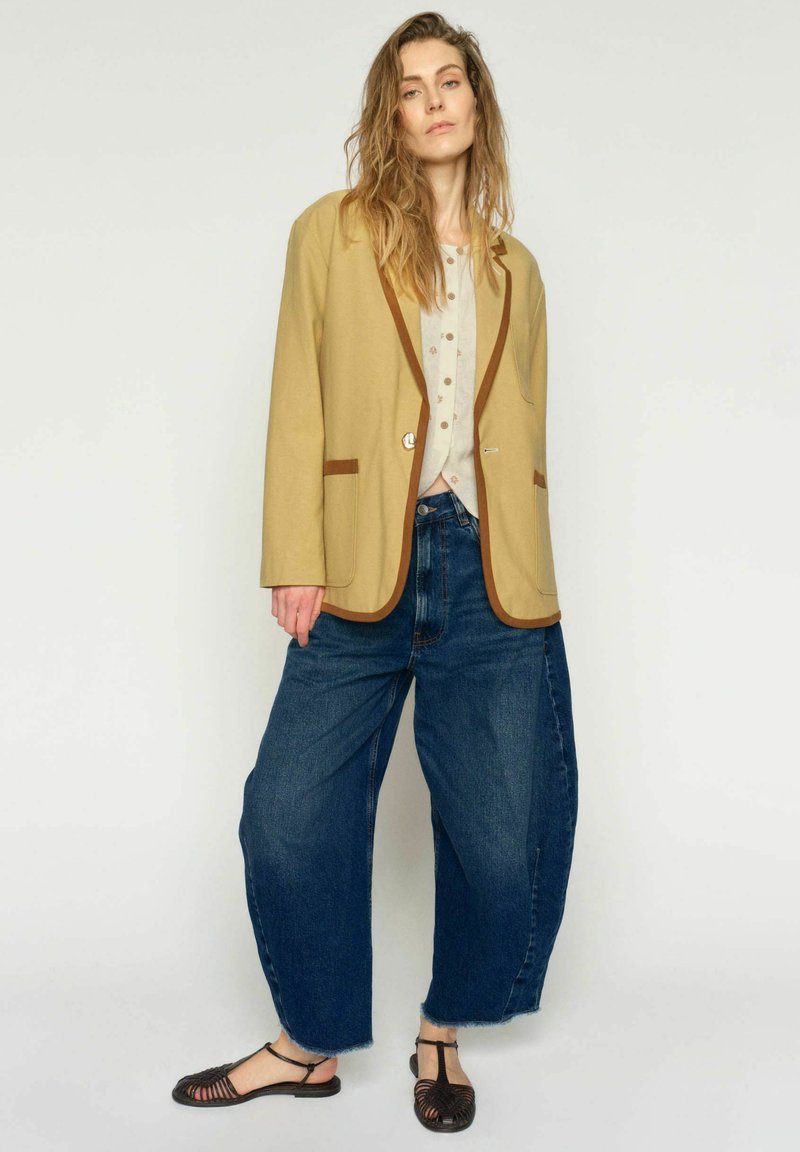 Nő bézs blézerben barna szegéllyel, világos mintás blúzzal, széles kék farmerral és fekete fonott szandállal, egy egyszínű háttér előtt állva.