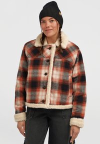 Vrouw met gevlochten haar, gekleed in een zwarte gebreide muts, een oranje- en zwarte geruite jas met witte sherpa voering, en zwarte broeken voor een witte achtergrond.