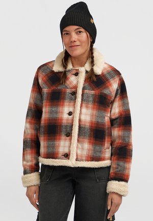 Donna con capelli intrecciati che indossa un berretto di maglia nero, una giacca a quadri arancione e nera con fodera in sherpa bianca e pantaloni neri, su uno sfondo bianco.