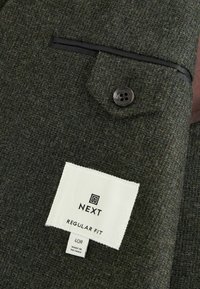 Tamnozelena teksturirana tkanina s jednim džepom na dugme. Ima oznaku s "NEXT", "REGULAR FIT" i veličinom 40R, napravljeno u Vijetnamu.