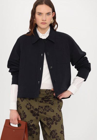 Navyblauwe cropped jas met een kraag, knoopsluiting aan de voorkant en een borstzak. Gedragen over een witte blouse en gecombineerd met gestreepte olijfgroene broek.