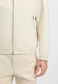 Giacca beige con zip, dal finish liscio e lucido, caratterizzata da un colletto alto, polsini elastici e una tasca laterale con una zip elegante.