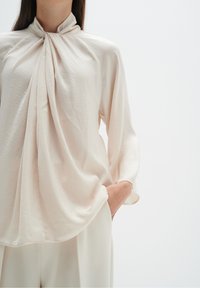 Blusa crema chiara con collo alto, dotata di tessuto drappeggiato sul davanti e maniche lunghe, realizzata in un materiale morbido e leggero.