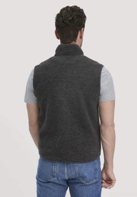 Dunkelgraue Fleece-Weste mit hohem Kragen, ärmellosem Design und weichem, strukturiertem Finish, über einem hellgrauen Hemd getragen.