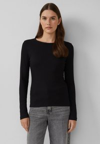 Zwarte langemouwen fitted top met een ronde hals en geribbelde textuur; voorzien van subtiele logo-details op de borst. Gecombineerd met grijze jeans.