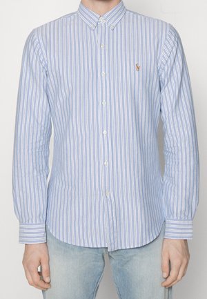 Man draagt een lichtblauw-wit verticale gestreept overhemd met knoopsluiting en een knoopkraag en lichtblauwe jeans, handen ontspannen langs de zij.