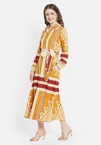 Abito camicia a maniche lunghe stampato in giallo, marrone e crema con motivi geometrici. Presenta una vita segnato da una cintura e polsini plissettati.