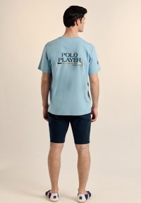 Una camiseta de algodón de color azul claro con un gráfico que dice "JUGADOR DE POLO" y un diseño de mazo en la parte posterior, combinada con pantalones cortos oscuros y zapatos informales.