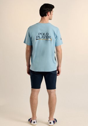 Una camiseta de algodón de color azul claro con un gráfico que dice "JUGADOR DE POLO" y un diseño de mazo en la parte posterior, combinada con pantalones cortos oscuros y zapatos informales.