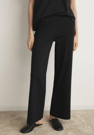 Czarne spodnie o szerokich nogawkach wykonane z miękkiego materiału, z elastycznym pasem, zestawione z luźną bluzką i prostymi czarnymi butami wsuwanymi.