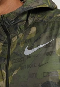 Close-up van een groene camouflagejas met een zwarte rits en herhaalde teksten "Just do it." en Nike-logo's, met een grote witte Nike swoosh op de borst.