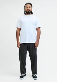 Moški z brado in dreadlocksi stoji, oblečen v belo majico, ohlapne črne jeans hlače in črne superge na enobarvni beli podlagi.