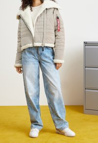 Krótka beżowa kurtka puchowa z obszyciem ze sztucznego owczej skóry, bocznymi kieszeniami na zamek i czerwoną metką. Połączona z jasnoniebieskimi szerokimi spodniami jeansowymi i białymi trampkami.