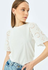 Hvid t-shirt med blonder i ærmerne og blomsterprint. Stoffet er blødt med en rund halsudskæring og en afslappet pasform. Kombinationen med blå denim.