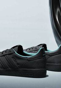 Paire de baskets noires Adidas Spezial avec doublure intérieure turquoise et logos Mercedes AMG sur le talon, présentées sur un fond clair.