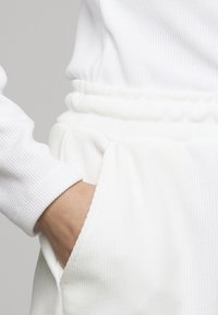 Χέρι στην τσέπη λευκού ριμπ πλεκτού παντελονιού με ελαστική μέση, συνδυασμένο με λευκό ριμπ πλεκτό μακρυμάνικο τοπ.