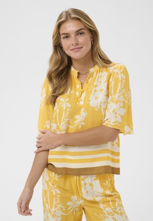 Kobieta o długich blond włosach, ubrana w żółtą, kwiecistą bluzkę i dopasowane spodnie, uśmiechająca się z założonymi ramionami na jasnym tle.
