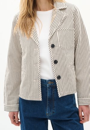 Frau trägt cremefarbenen und schwarzen, vertikal gestreiften Blazer mit schwarzen Knöpfen, weißes Oberteil und blaue Jeans, Hände entspannt an den Seiten.