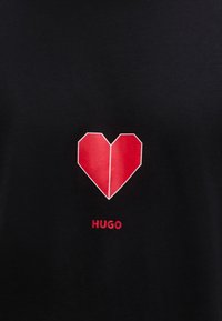 Μαύρο μπλουζάκι με κόκκινη γεωμετρική καρδιά, περιγράμμενη σε λευκό, με τη λέξη "HUGO" τυπωμένη σε κόκκινο κάτω από την καρδιά.