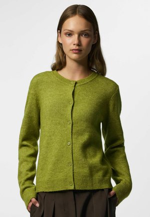 Jeune femme portant un cardigan vert boutonné et un pantalon foncé, debout contre un fond clair uni, les mains dans les poches.