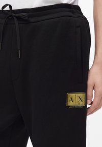Черни спортни панталони Armani Exchange с връзка на талията и златен лого етикет на дясното бедро, носени с бяла риза.