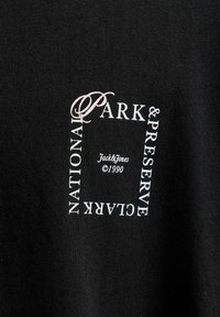 Melnā T-krekls ar centrālo balto un rozā grafiku, kas uzrakstīts "NATIONAL PARK & PRESERVE CLARK," ar "Jack & Jones ©1990" zem tā.