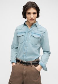 Νεαρό άτομο που φοράει ανοιχτό γαλάζιο τζιν πουκάμισο με τσέπες στο στήθος και καφέ παντελόνι, στέκεται με τα χέρια στις τσέπες μπροστά σε απλό φόντο.