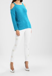 Pull en tricot turquoise avec un design épaule dénudée, associé à un pantalon blanc ajusté avec des accents de clous dorés et des talons argentés brillants.