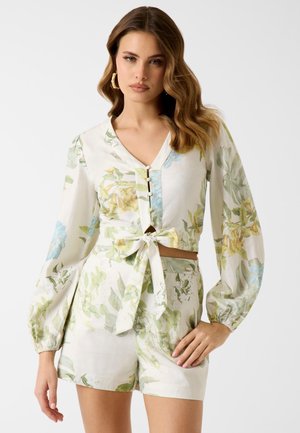 Femme portant un chemisier blanc à motif floral avec col en V, boutons et manches longues bouffantes, associé à un short taille haute assorti avec ceinture à nouer.