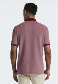Homme portant un polo texturé bordeaux et blanc avec un col et des poignets de manches rouge foncé, se tenant de dos, en pantalon beige.