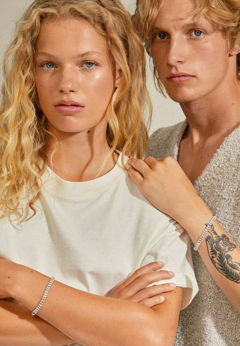 Due modelle con capelli biondi mossi. Una indossa una maglietta beige chiaro, l'altra un maglione grigio con texture. Entrambe portano bracciali in argento.