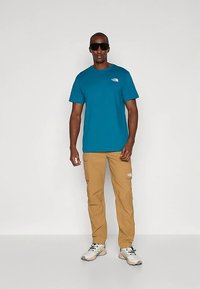 Moški v modri kratki majici s kratkimi rokavi, bež hlačah s številnimi žepi, belih pohodnih čevljih in črnih velikih sončnih očalih stoji pred enobarvnim ozadjem.