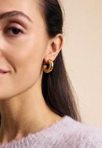 Goldene Creolen mit einer glatten, polierten Oberfläche. Kreisförmiges Design mit einem leichten Schwung, im Ohr positioniert vor einem neutralen Hintergrund.