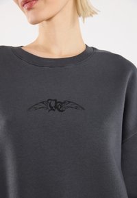 Felpa grigia scura con colletto tondo. Presenta una grafica nera con ali e lettere sul petto. Texture morbida, vestibilità rilassata.