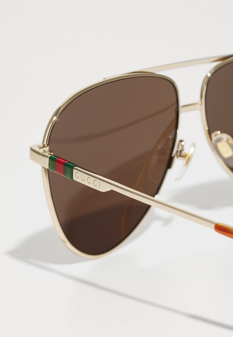 Occhiali da sole aviator in metallo dorato con lenti marroni. La asticella presenta un design a strisce verde e rossa con "Gucci" inciso sul lato.