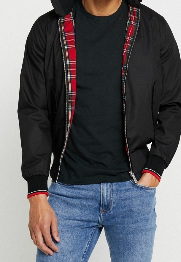 Man med en svart zip-up jacka med röd rutigt foder, svart t-shirt och blå jeans, med en hand i fickan och den andra avslappnad.