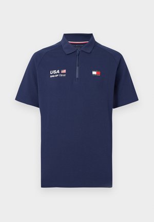 Polo de manga corta azul marino con media cremallera, con texto USA SailGP Team, bandera y logo de Tommy Hilfiger en el pecho.