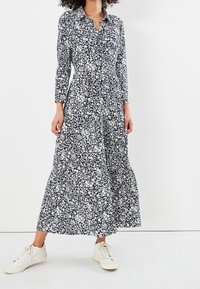 Dlouhé, tmavě modré šaty s bílým květinovým vzorem, zapínání na knoflíky vpředu, dlouhé rukávy a rozšířená sukně, kombinované s bílými teniskami.