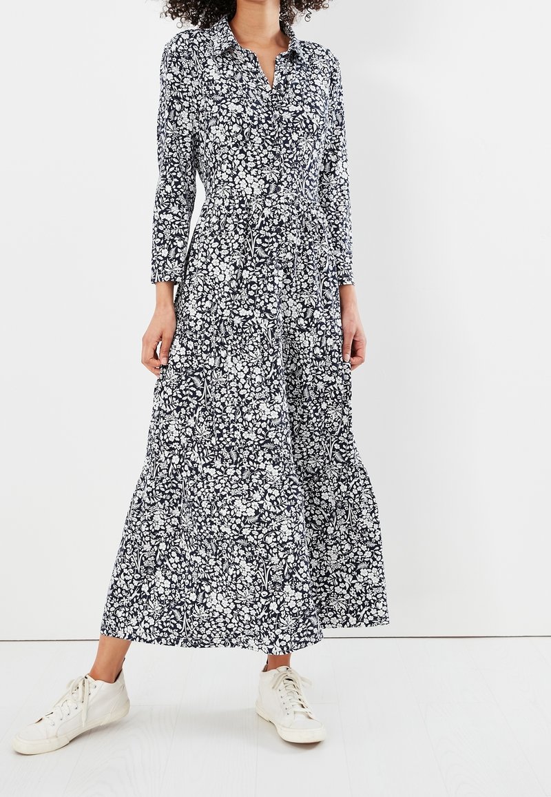 Pitkä tummansininen mekko valkoisella kukkakuviolla, nappikiinnitys edessä, pitkät hihat ja levenevä hameosa, yhdistettynä valkoisiin lenkkareihin.