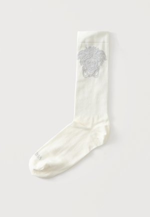 Weißer gerippter Socken mit grauem Medusenhaupt-Design nahe dem oberen Bündchen, flach liegend auf hellem Hintergrund.