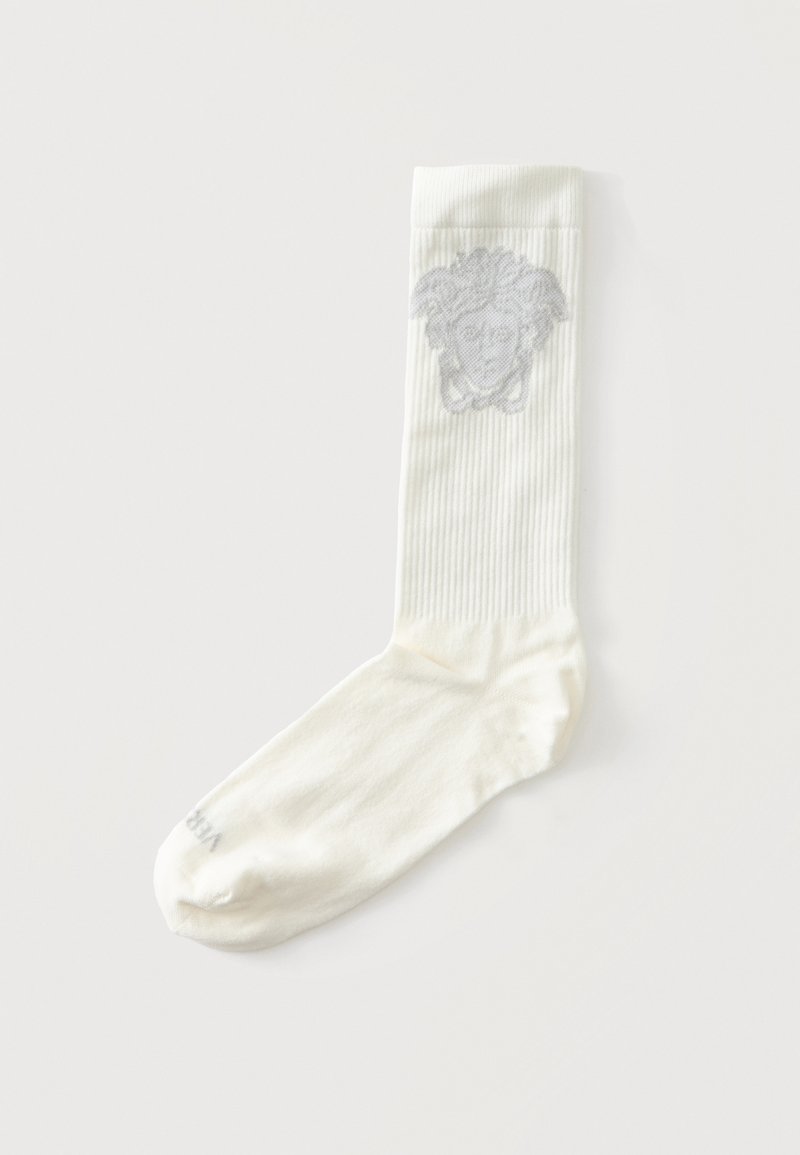Chaussette blanche côtelée avec un dessin gris de tête de Méduse près du bord supérieur, posée à plat sur un fond clair.
