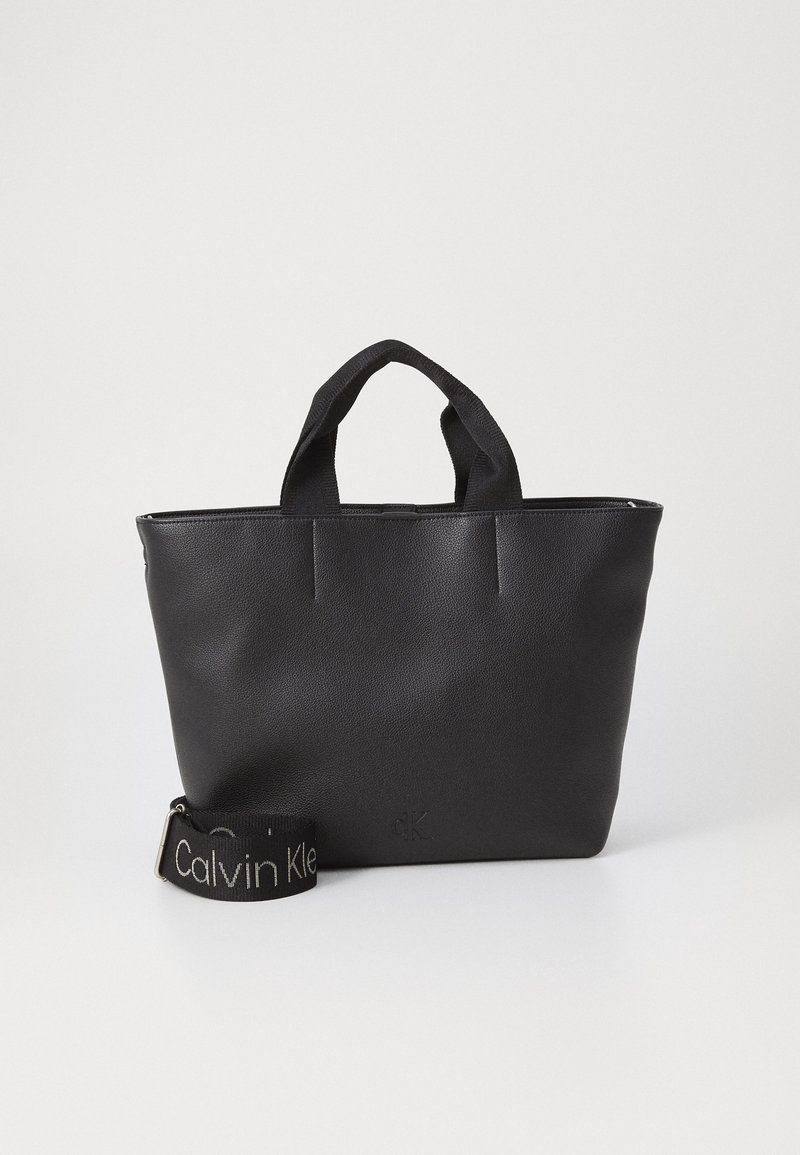 Sac en cuir noir avec double poignée supérieure, texture lisse et détail de logo. Comprend une sangle amovible avec la marque "Calvin Klein".