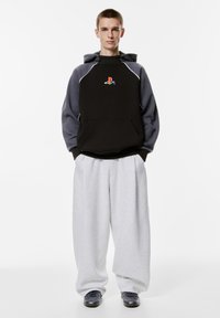 Czarna bluza z kapturem z szarymi rękawami, z kolorowym logo PlayStation. W zestawieniu z luźnymi szarymi spodniami dresowymi i ciemnymi butami.