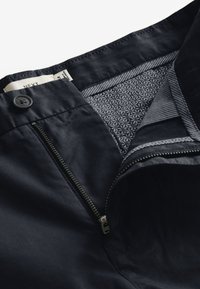 Dunkelmarineblaue Hose mit einem Reißverschluss und Knopfverschluss vorne, sowie einem strukturierten inneren Taillenbund im grauen geometrischen Muster.