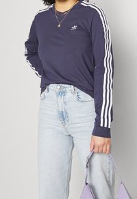 Námornická košile s dlouhým rukávem a bílými pruhy na rukávech a malým logem. Kombinováno se světle modrými džíny s vysokým pasem a fialovou kabelkou.