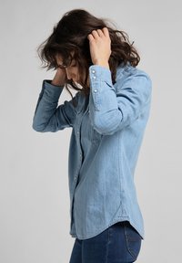Donna con capelli ricci scuri, indossa una camicia di jeans blu chiaro e jeans. Tiene la testa con entrambe le mani e guarda verso il basso, su uno sfondo grigio uniforme.