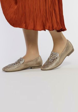 Zapatos slip-on de oro tejido con una superficie texturada y diseño recortado en la punta. Suela plana, acompañados de una falda plisada naranja.