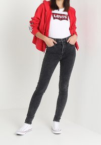Sudadera roja con cremallera, camiseta blanca con el logo de Levi's, pantalones skinny negros y zapatillas blancas. Modelo posado sobre un fondo claro.