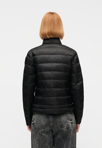 Personne aux cheveux courts roux portant une veste noire matelassée et un jean noir délavé, debout face à un fond blanc uni.