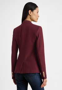 Vīna sarkana žakete no teksturētas auduma ar pieguļošu dizainu, ar zelta pogu akcentiem piedurknēs un šķēlumu aizmugurē.